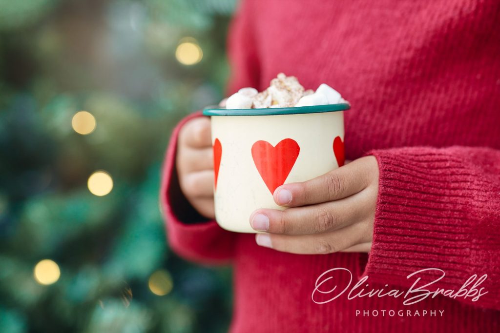child's hands holding scandi style cup of hot chocolate in front of christmas tree